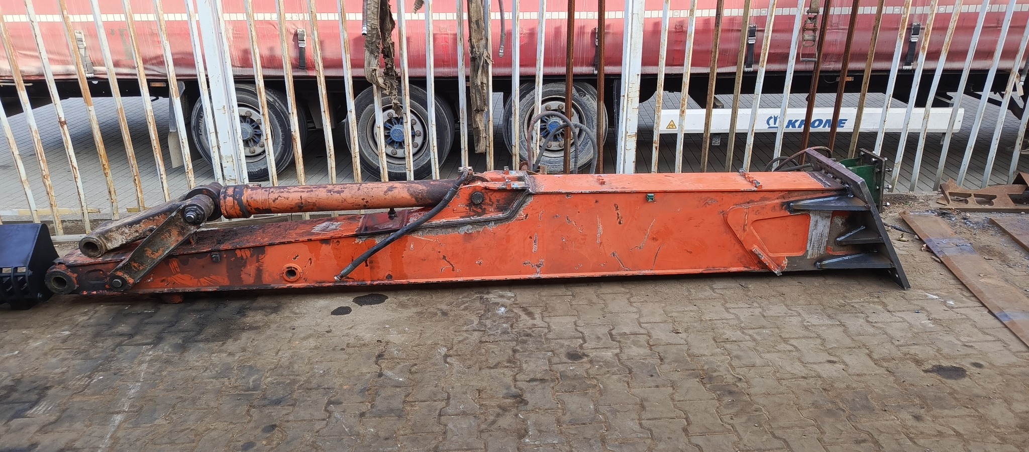 Pomarańczowe ramię koparki z hydraulicznym siłownikiem, leżące na brukowanej powierzchni, z widocznym fragmentem naczepy ciężarówki w tle.