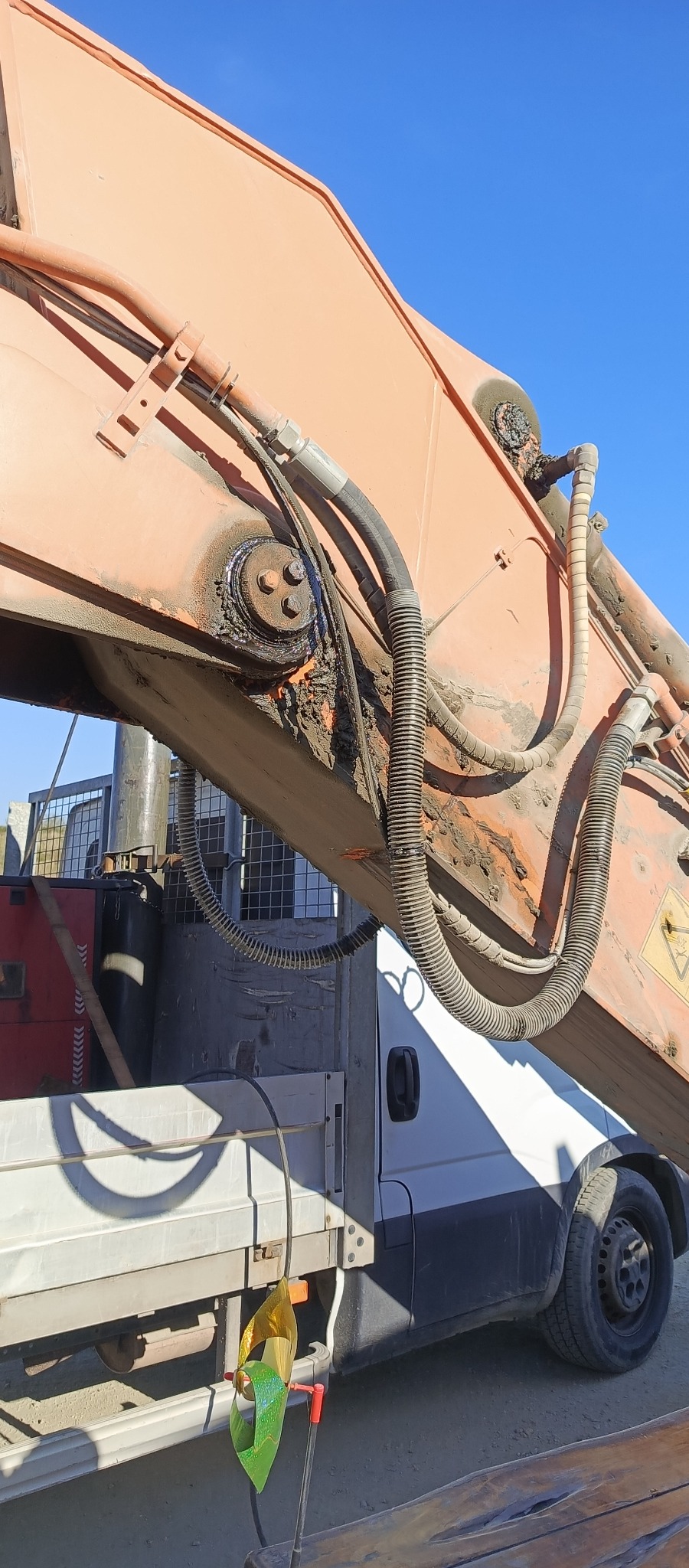 Pomarańczowe ramię dźwigu hydraulicznego zamontowane na pojeździe dostawczym, widoczne przewody hydrauliczne, złącza i elementy mocujące, częściowo zabrudzone ziemią, na tle błękitnego nieba.