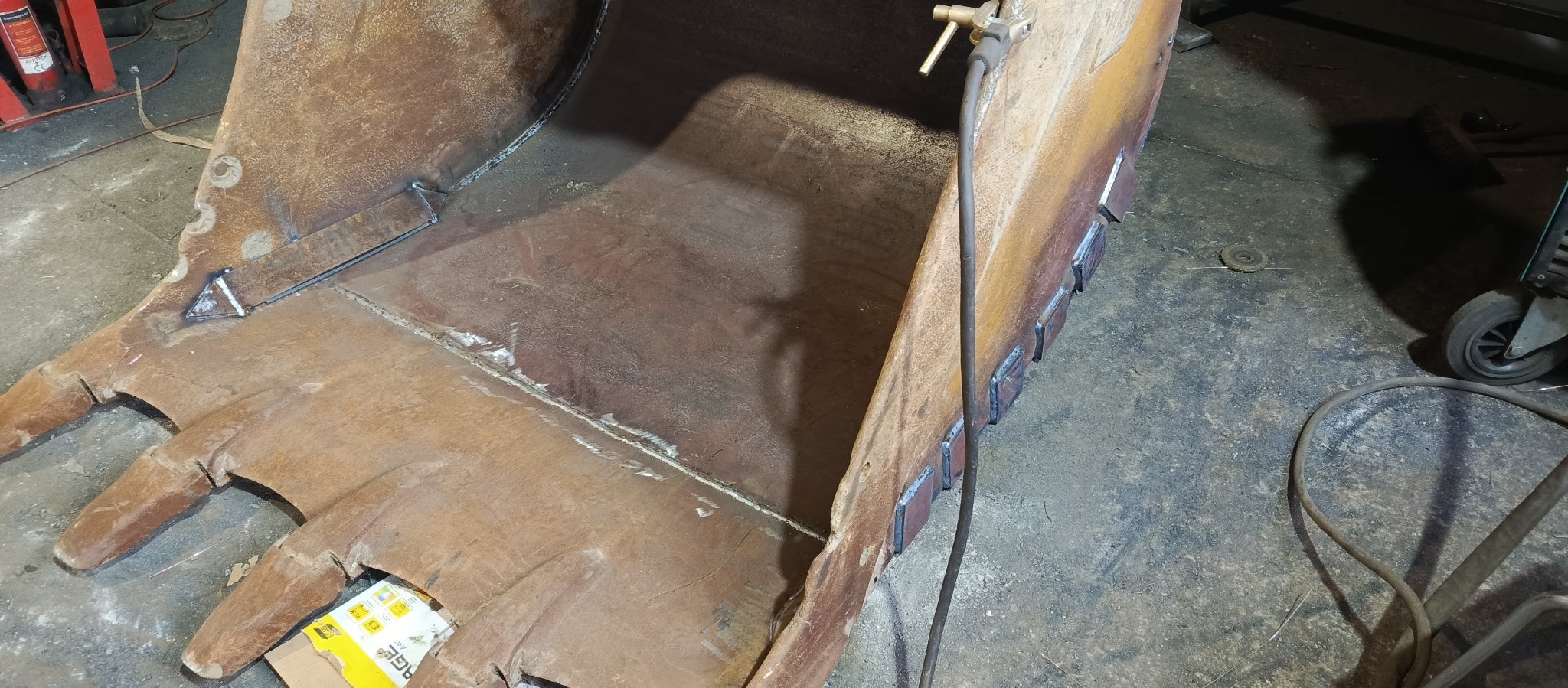 Wnętrze łyżki koparki z widocznymi świeżymi spawami wzmacniającymi konstrukcję, uchwyty do mocowania oraz spawarka w tle.