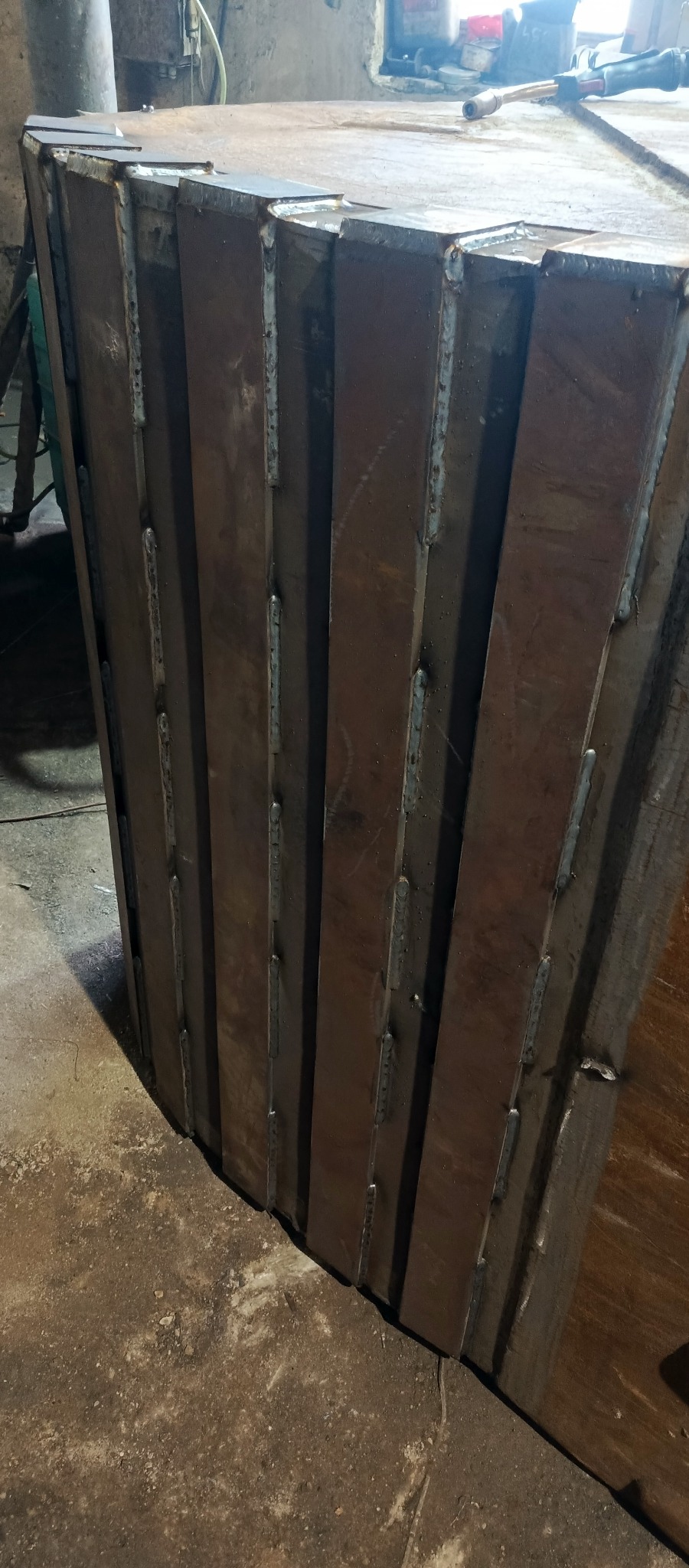 Fragment konstrukcji stalowej z widocznymi spawami, wykonanej z kilku pionowych elementów połączonych ze sobą, w warsztacie.