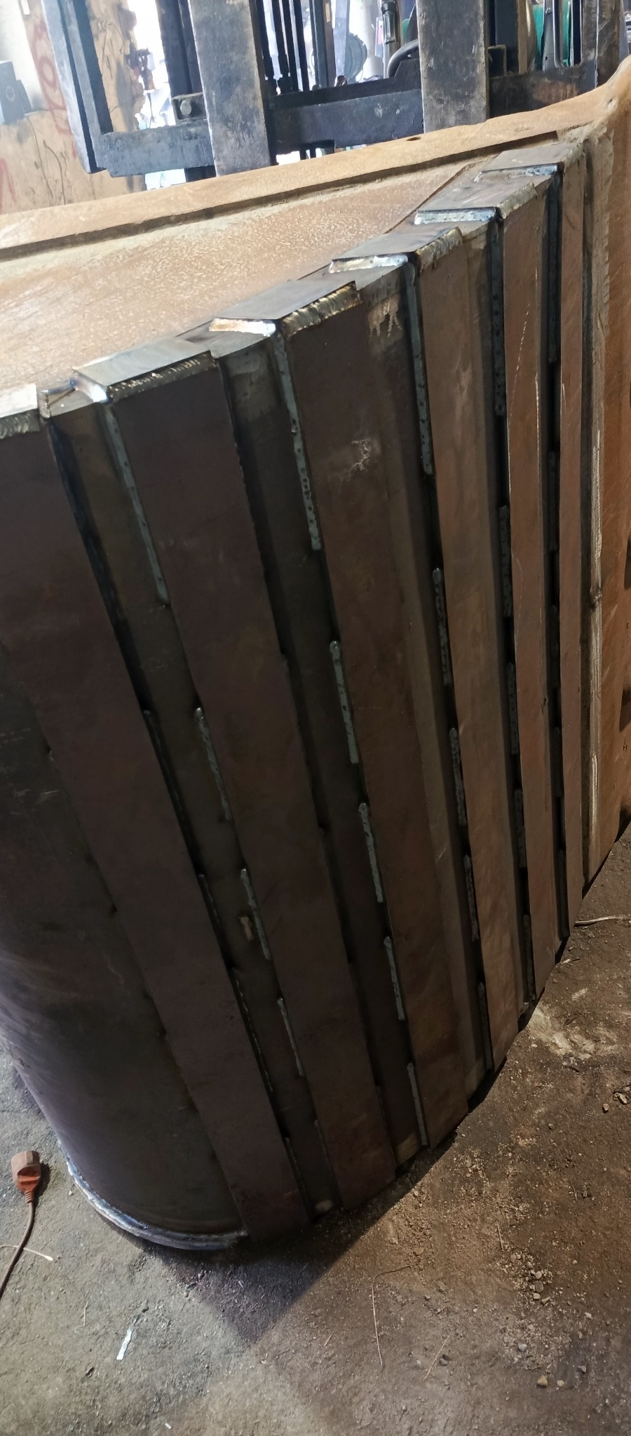 Fragment konstrukcji stalowej z widocznymi spawami, wykonanej z segmentów, w tle widoczny wózek widłowy.