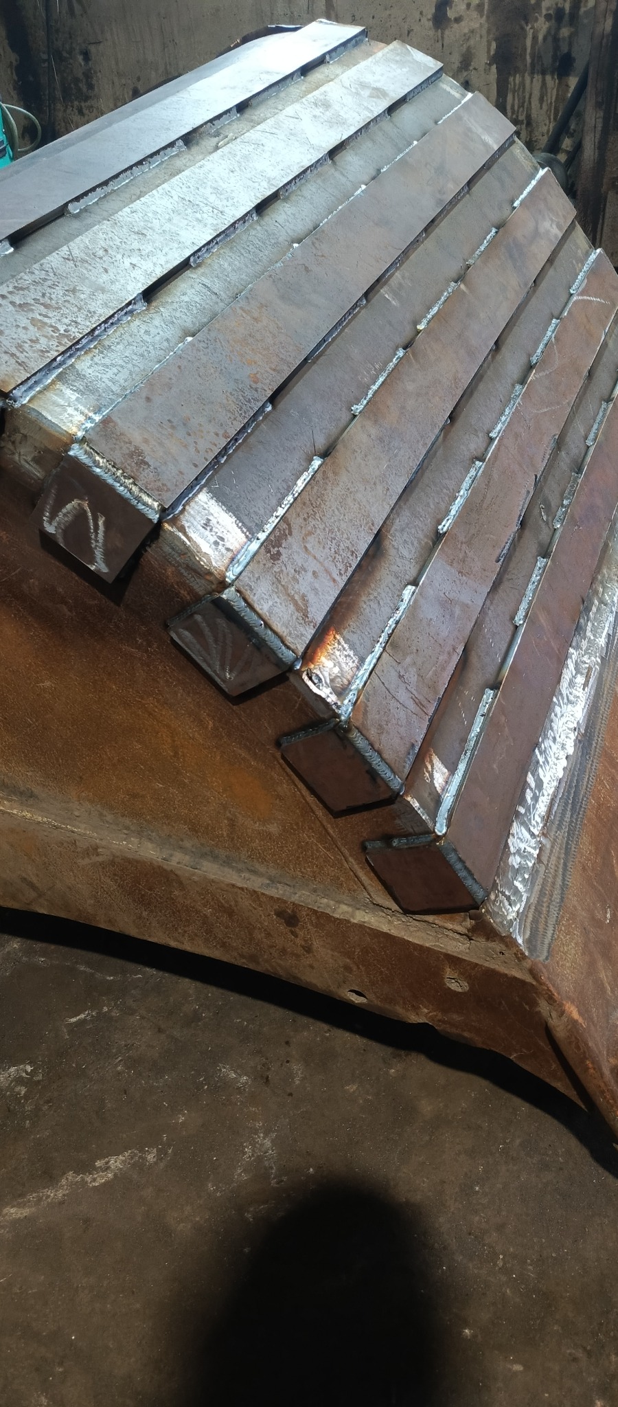 Fragment konstrukcji stalowej z przyspawanymi elementami, widoczne spawy i surowy wygląd metalu.