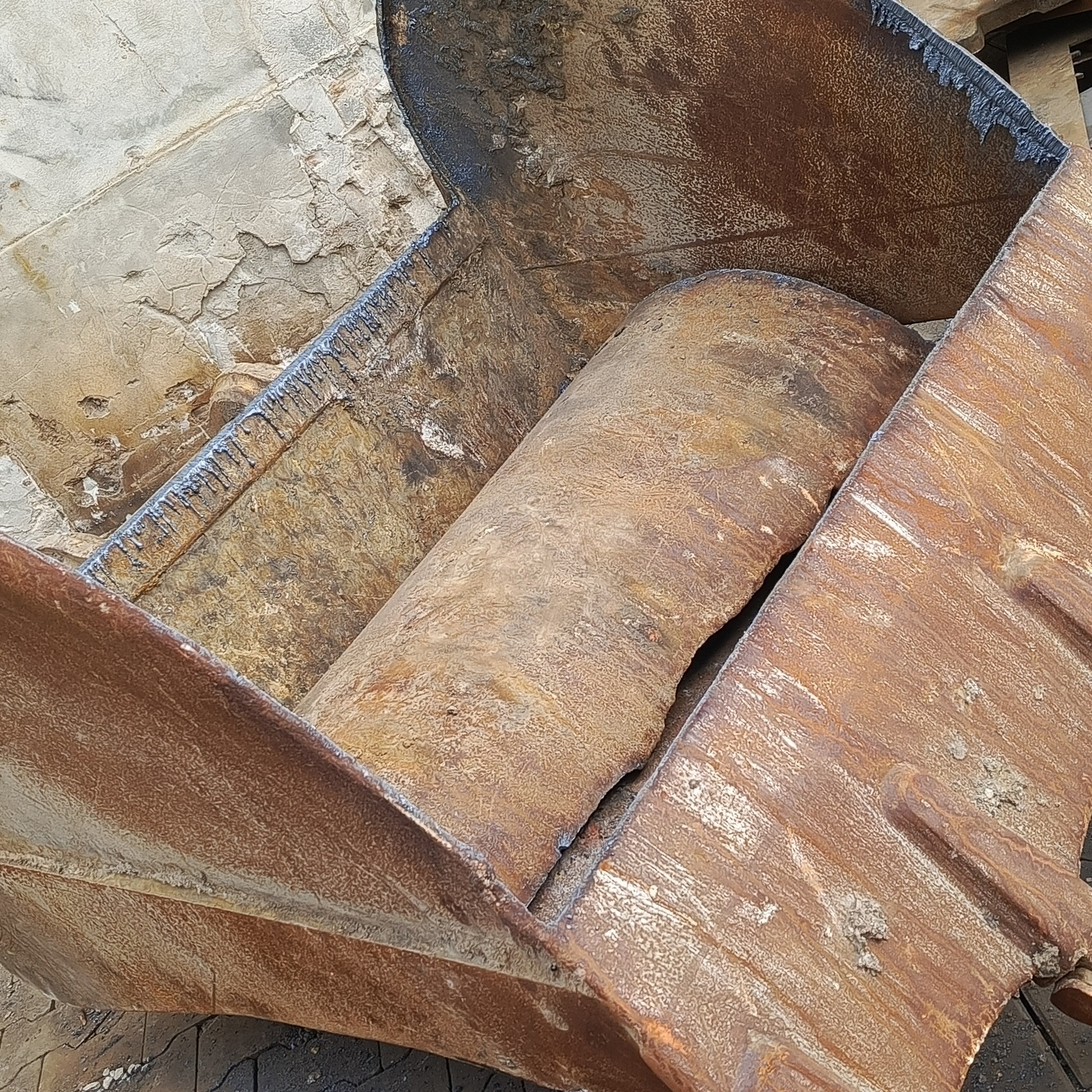 Wnętrze brudnej, zardzewiałej łyżki koparki z fragmentem ziemi w środku, widoczne ślady użytkowania i zniszczenia.