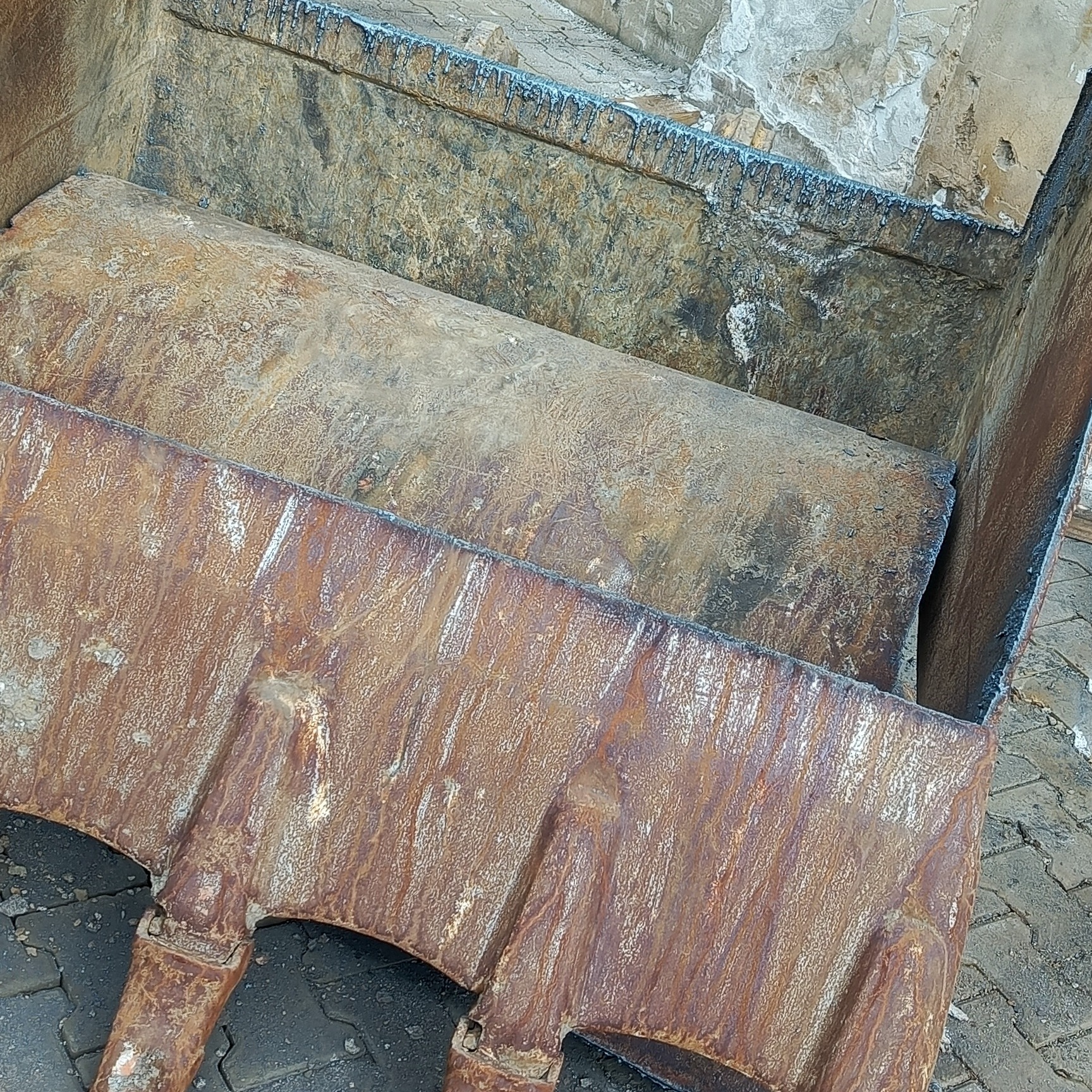 Szczegółowe ujęcie zardzewiałej, metalowej łyżki koparki z widocznymi śladami spawania i użytkowania, leżącej na brukowanej powierzchni.