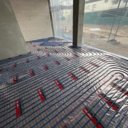 Węże ogrzewania podłogowego rozłożone na folii z siatką w surowym wnętrzu budynku w Ostrołęce, widok z góry na instalację przed wylaniem posadzki.