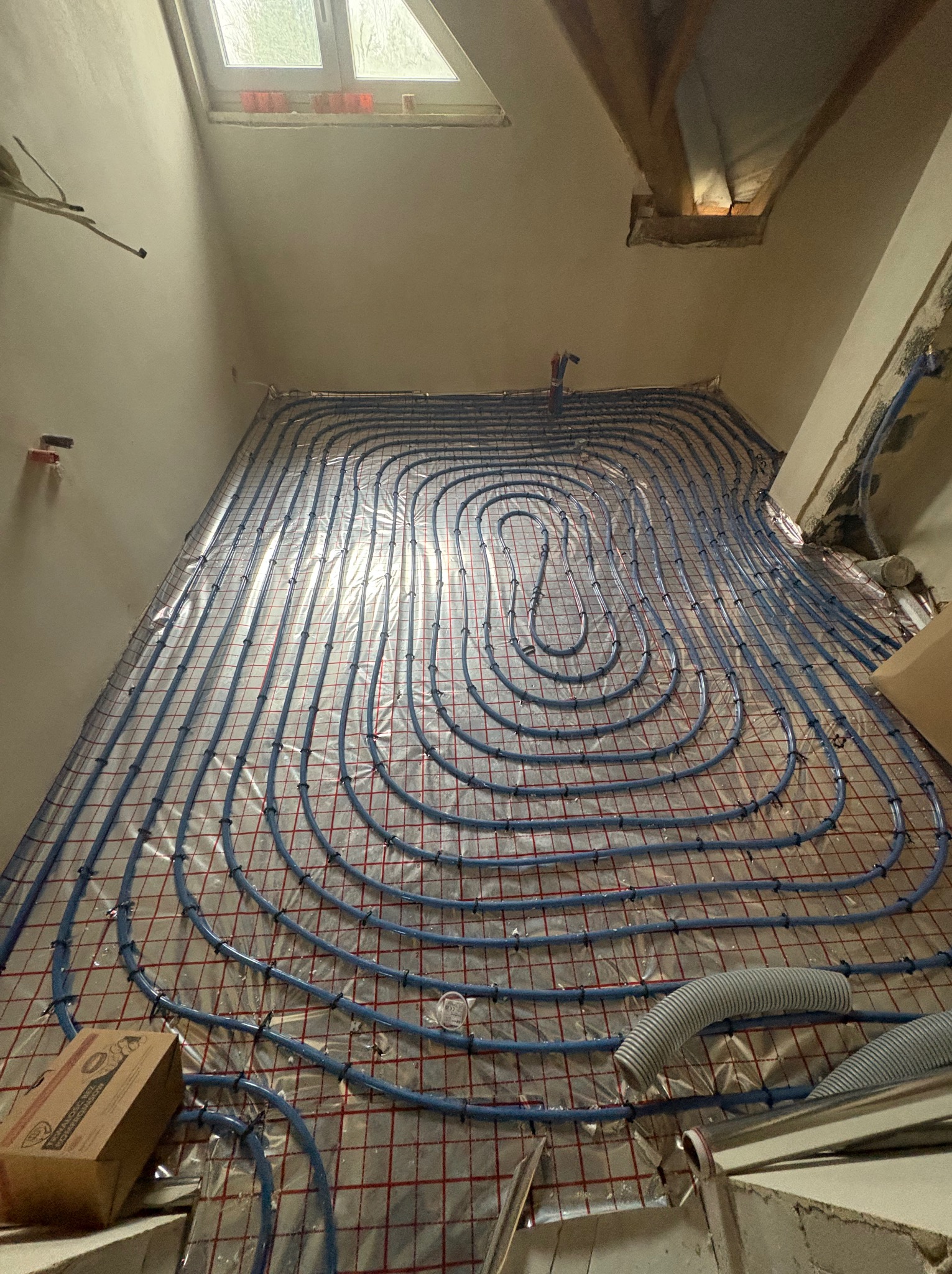Niedokończona instalacja ogrzewania podłogowego z niebieskich rurek na folii z czerwoną siatką, widoczne fragmenty ścian i okno dachowe w surowym stanie.