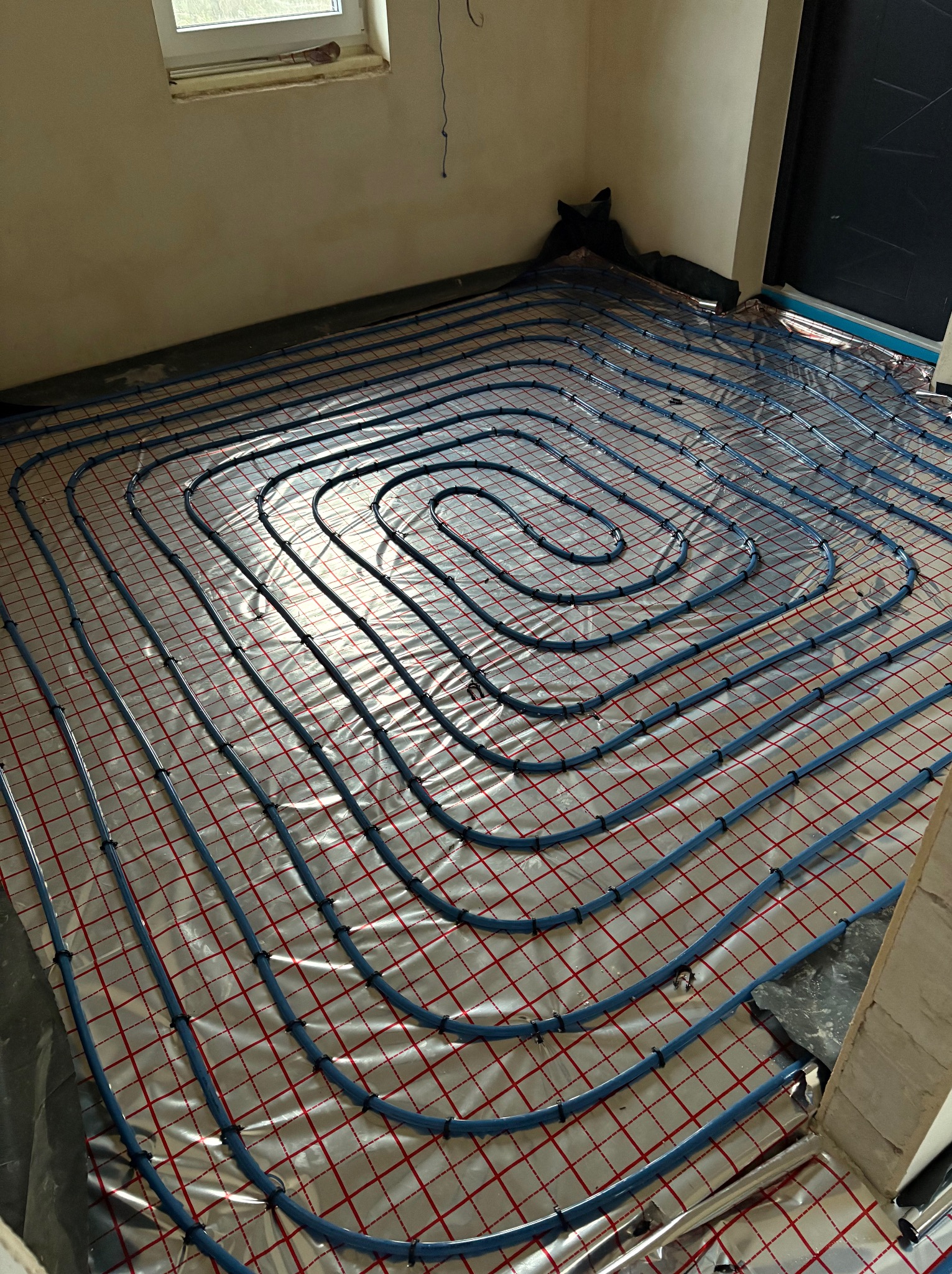 Niebieskie rurki ogrzewania podłogowego ułożone spiralnie na izolacji z czerwonymi kwadratami, widok z góry na surową podłogę przed wylewką.
