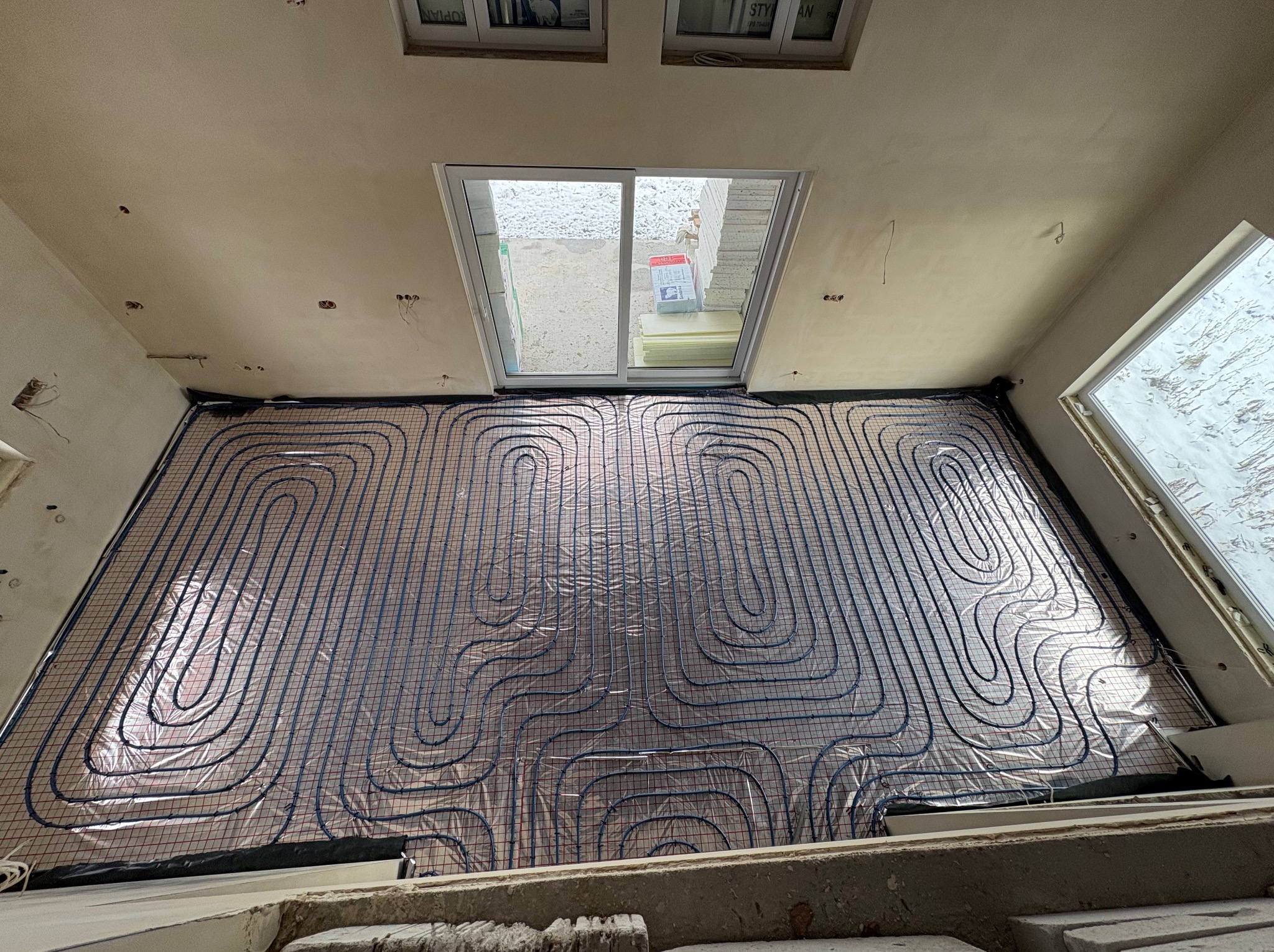 Niedokończony montaż ogrzewania podłogowego z rurkami ułożonymi na siatce, widoczne okno dachowe i materiały budowlane na zewnątrz.