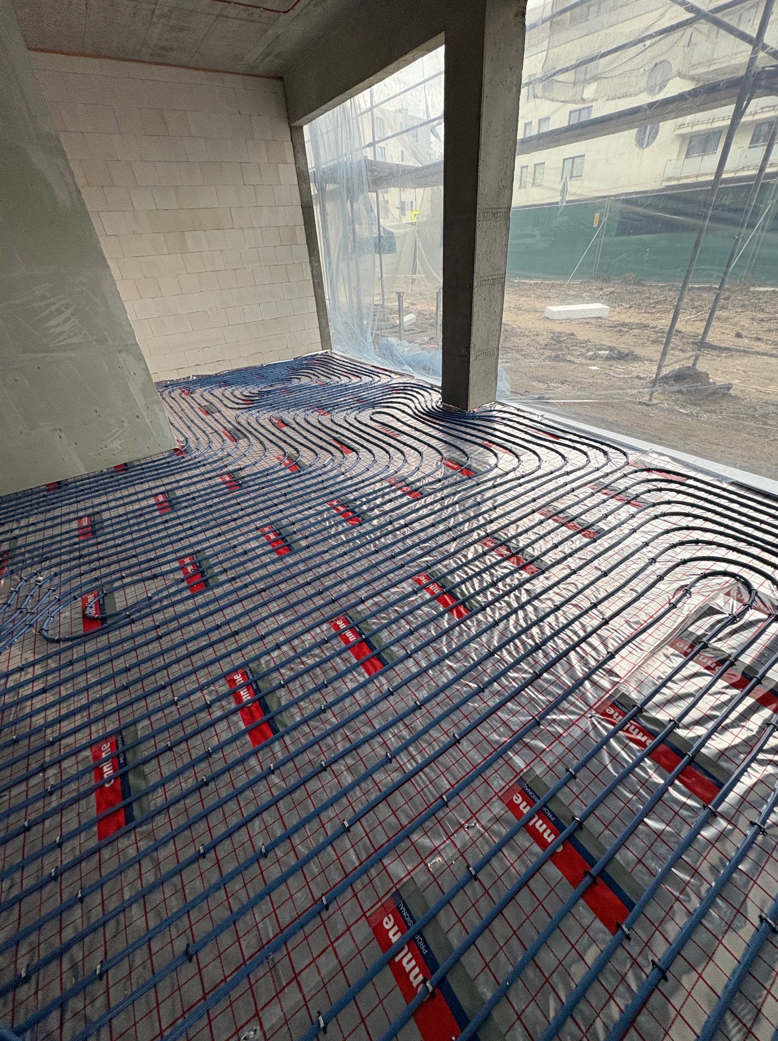 Węże ogrzewania podłogowego rozłożone na folii z siatką w surowym wnętrzu budynku w Ostrołęce, widok z góry na instalację przed wylaniem posadzki.
