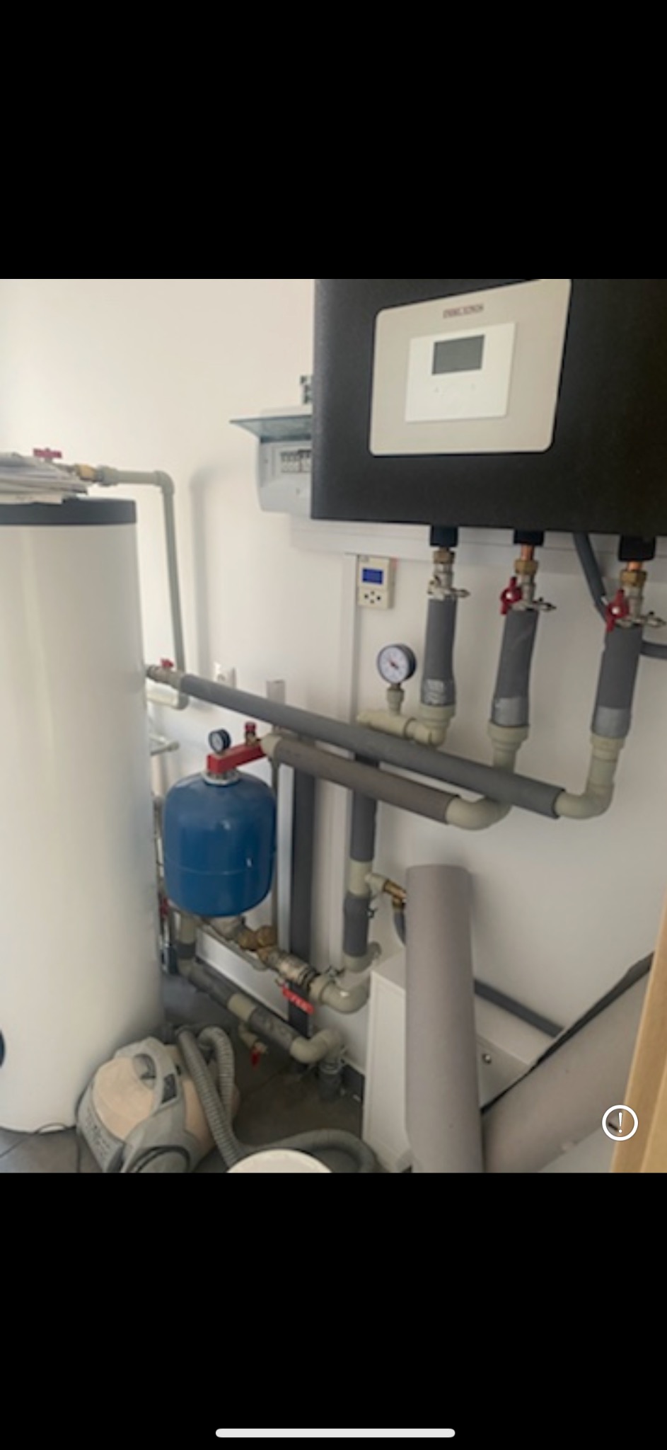Instalacja grzewcza z zasobnikiem ciepłej wody, naczyniem przeponowym i piecem ze sterownikiem elektronicznym, widoczne rury izolowane termicznie i manometry, obok odkurzacz.