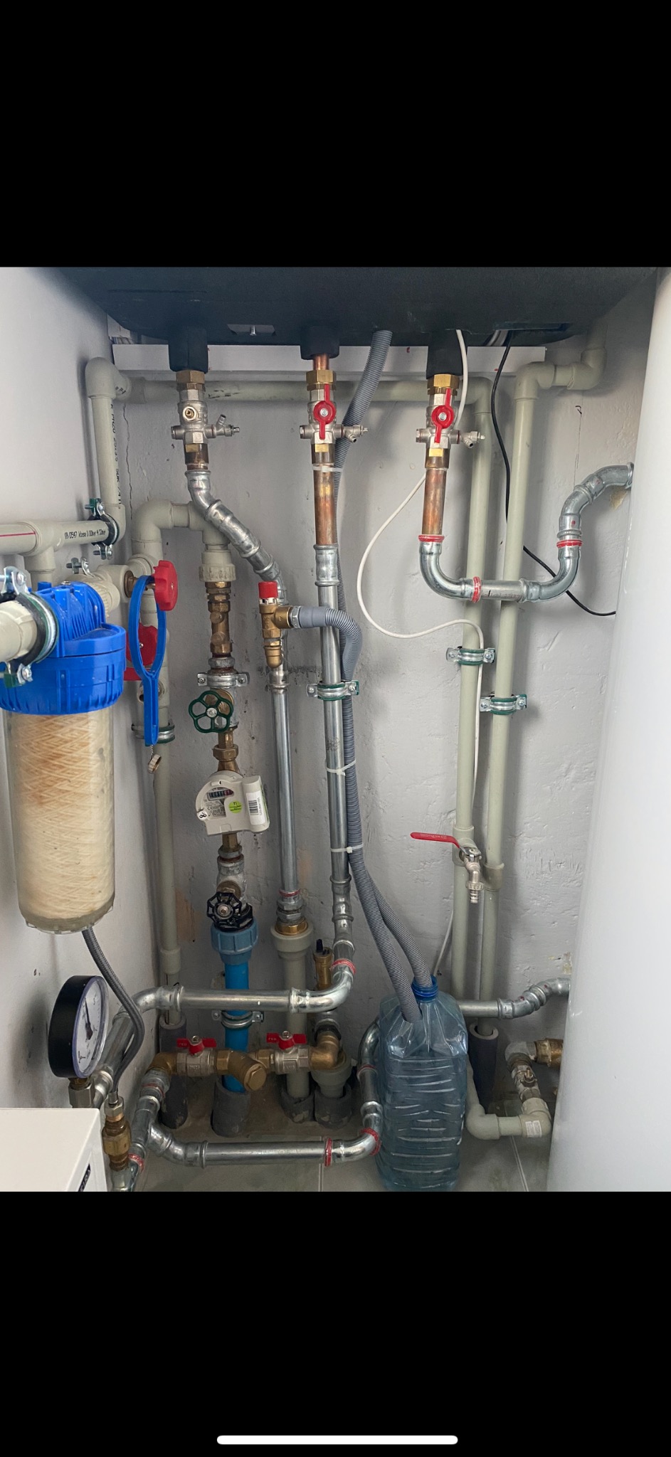 Złożony system rur i zaworów wodnych z manometrem, filtrem wody z widocznym osadem, oraz butelką z wodą podłączoną do instalacji, całość na tle białej ściany.