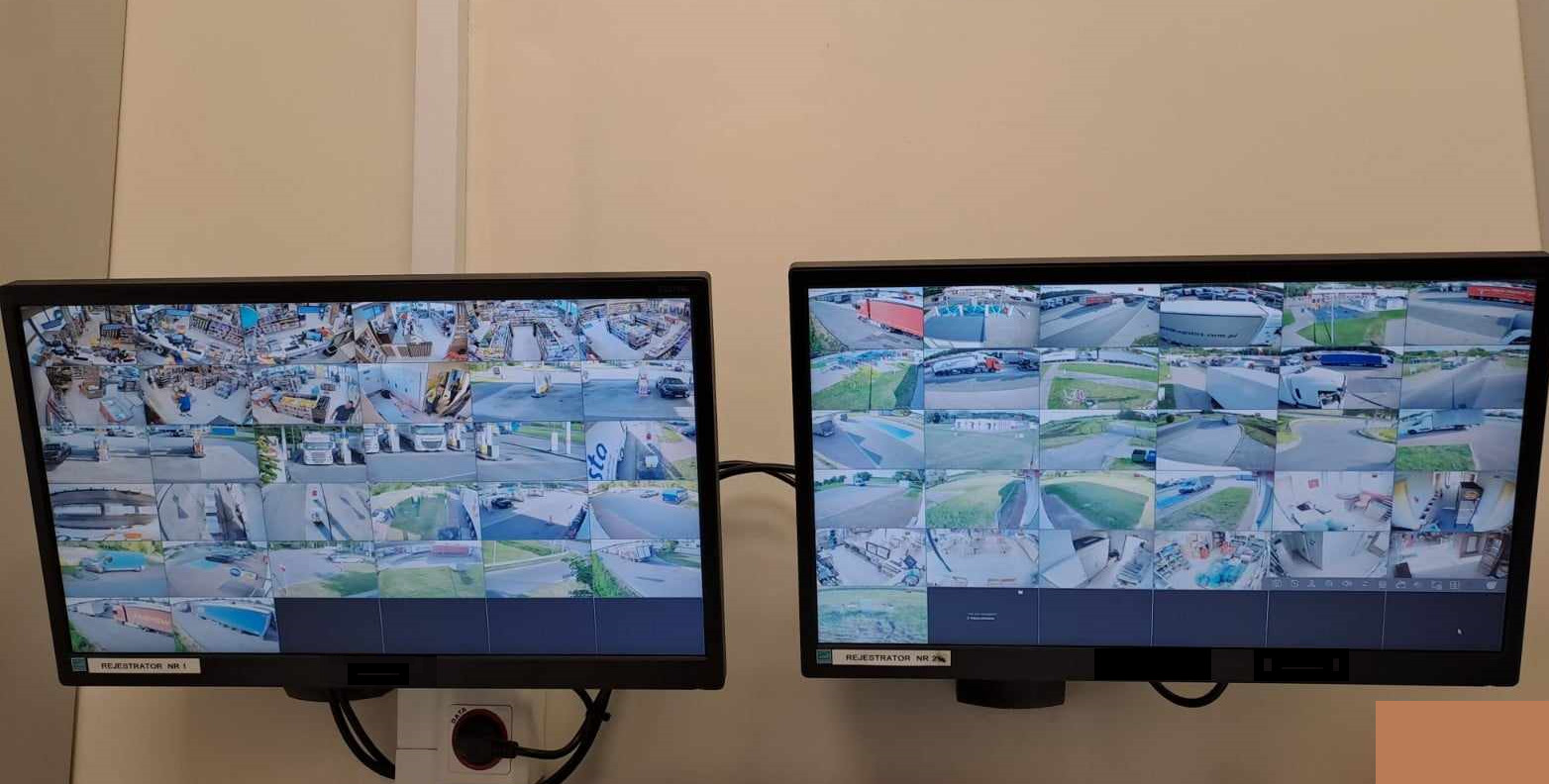 Dwa monitory LCD wyświetlające obraz z kamer przemysłowych; lewy monitor pokazuje wnętrze sklepu, prawy - parking z pojazdami ciężarowymi.
