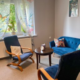 Mateusz Kur Psychoterapia i Edukacja - Przytulny pokój z niebieską sofą, dwoma fotelami z drewnianymi podłokietnikami i okrągłym stolikiem. Okno z zasłonami w motyle i rośliny doniczkowe. Na ścianie wiszą obrazy w jasnych ramach.