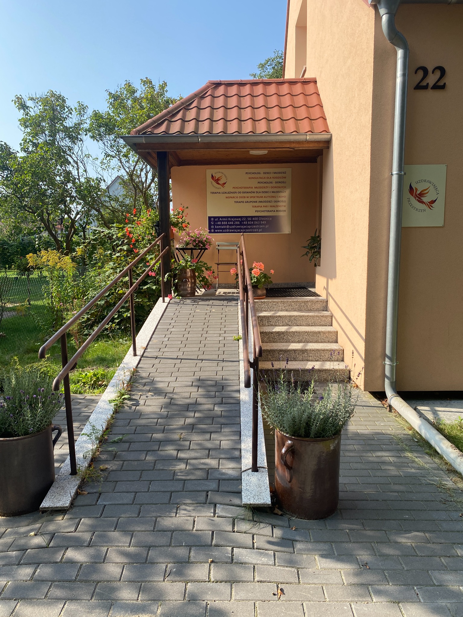 Wejście do budynku z rampą i schodami, tablica informacyjna z listą usług, donice z lawendą po bokach, numer budynku 22.