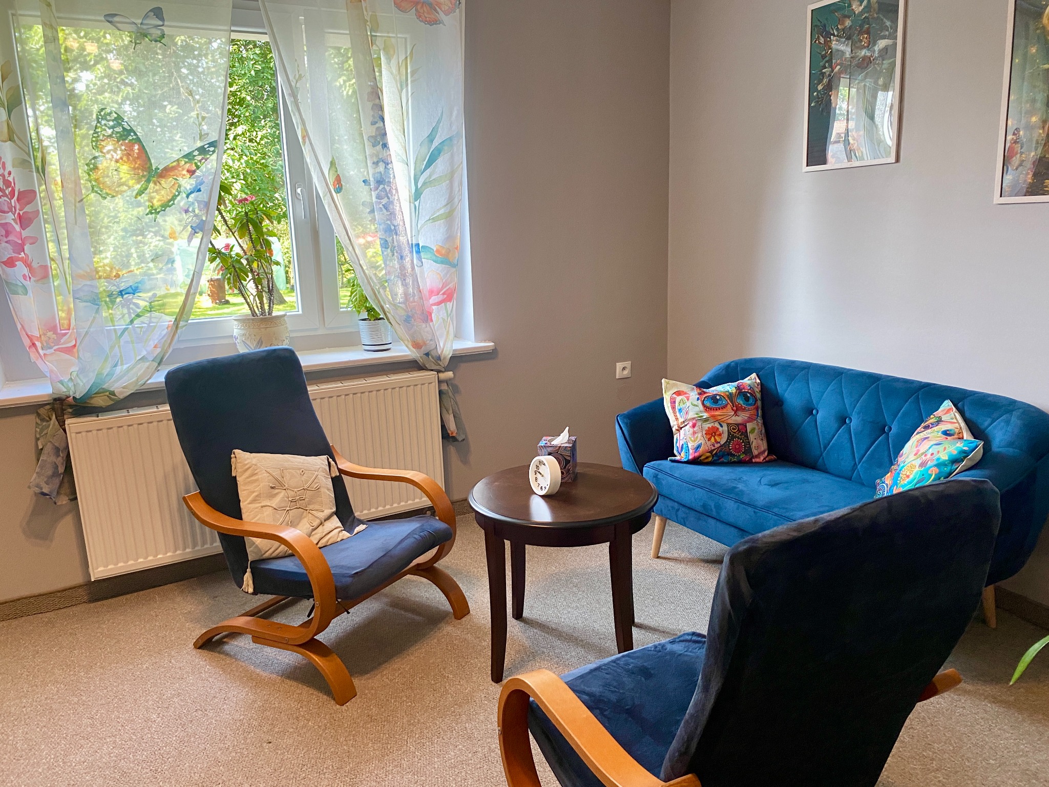 Przytulny pokój z niebieską sofą, dwoma fotelami z drewnianymi podłokietnikami i okrągłym stolikiem. Okno z zasłonami w motyle i rośliny doniczkowe. Na ścianie wiszą obrazy w jasnych ramach.