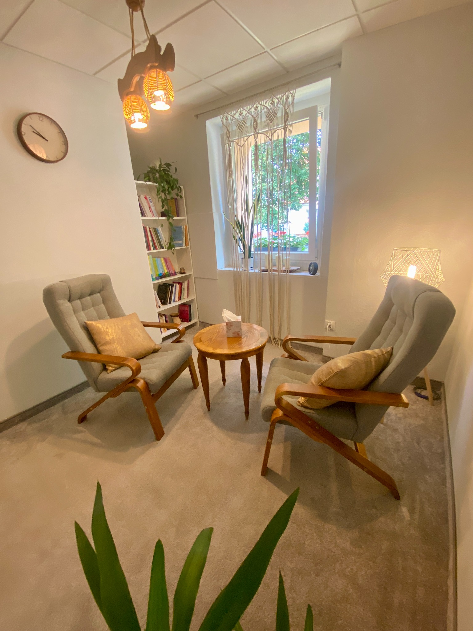 Jasne wnętrze gabinetu z dwoma fotelami, drewnianym stolikiem, lampą, zegarem na ścianie i regałem z książkami, doświetlone naturalnym światłem z okna z delikatną firanką.