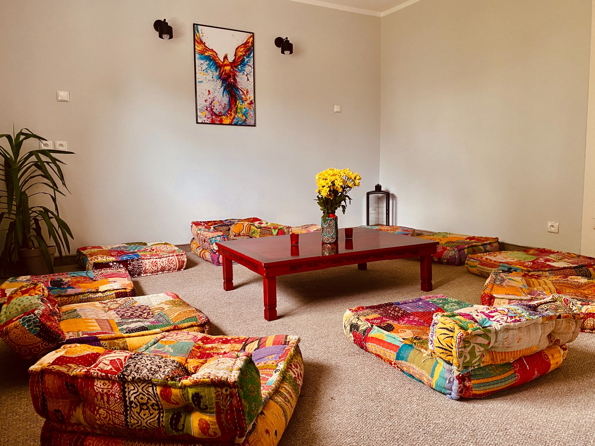 Pokój z kolorowymi pufami do siedzenia ułożonymi wokół niskiego, czerwonego stolika z bukietem żółtych kwiatów, na ścianie wisi obraz z motywem feniksa.
