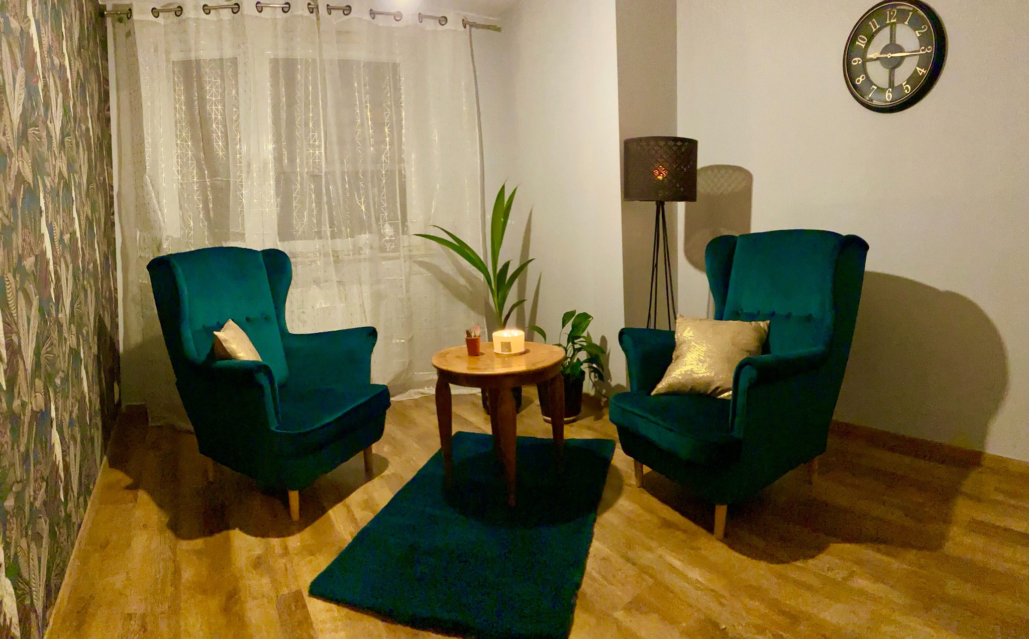 Pokój z dwoma zielonymi fotelami typu uszak, małym drewnianym stolikiem, lampą stojącą, zegarem na ścianie i roślinami doniczkowymi, tworzący przytulną przestrzeń do rozmowy.