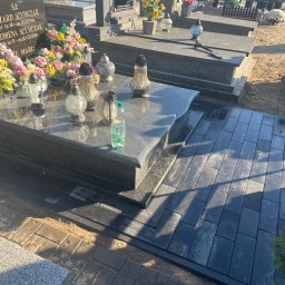 Max Bruk - Ułożona kostka brukowa wokół nagrobka z ciemnego granitu, widoczne kwiaty i znicze na płycie nagrobnej, butelka wody, częściowo widoczna stara kostka brukowa na obrzeżach.