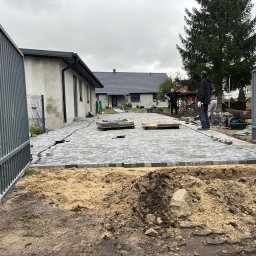 Max Bruk - Kładzenie szarej kostki brukowej na podwórku domu jednorodzinnego, widoczni pracownicy w trakcie robót, palety z kostką i narzędzia, w tle drzewo iglaste i sąsiedni budynek.