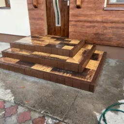 Trzy stopnie wejściowe wyłożone brązowo-żółtą kostką, prowadzące do drewnianych drzwi z szybą, elewacja budynku z drewnopodobnych paneli, fragment mokrego chodnika i zielonego węża ogrodowego...