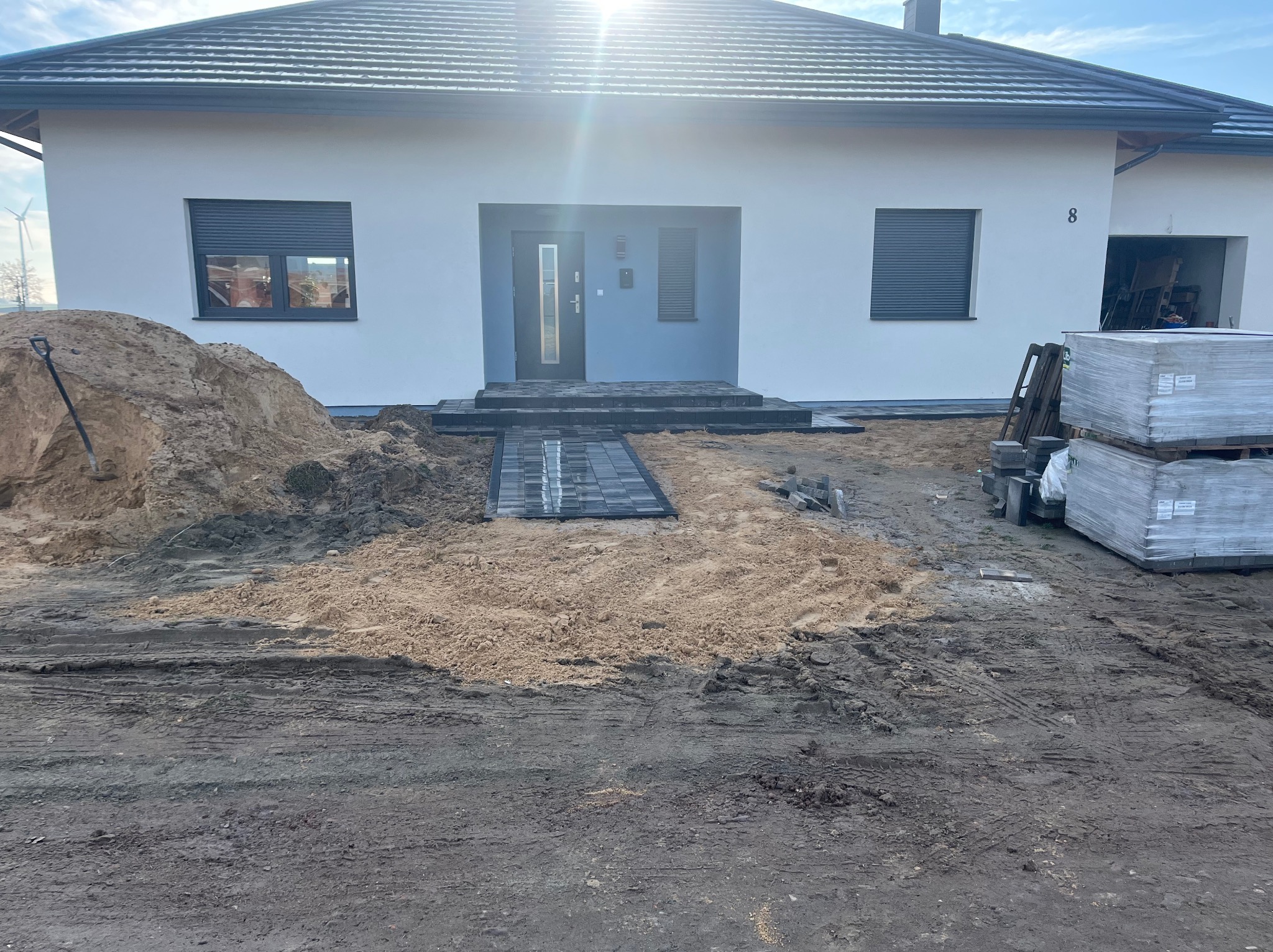 Wjazd do domu jednorodzinnego w trakcie prac brukarskich, widoczny fragment ułożonej ścieżki z kostki grafitowej, hałda piasku i paleta z kostką brukową. W tle biała elewacja budynku z szarymi...