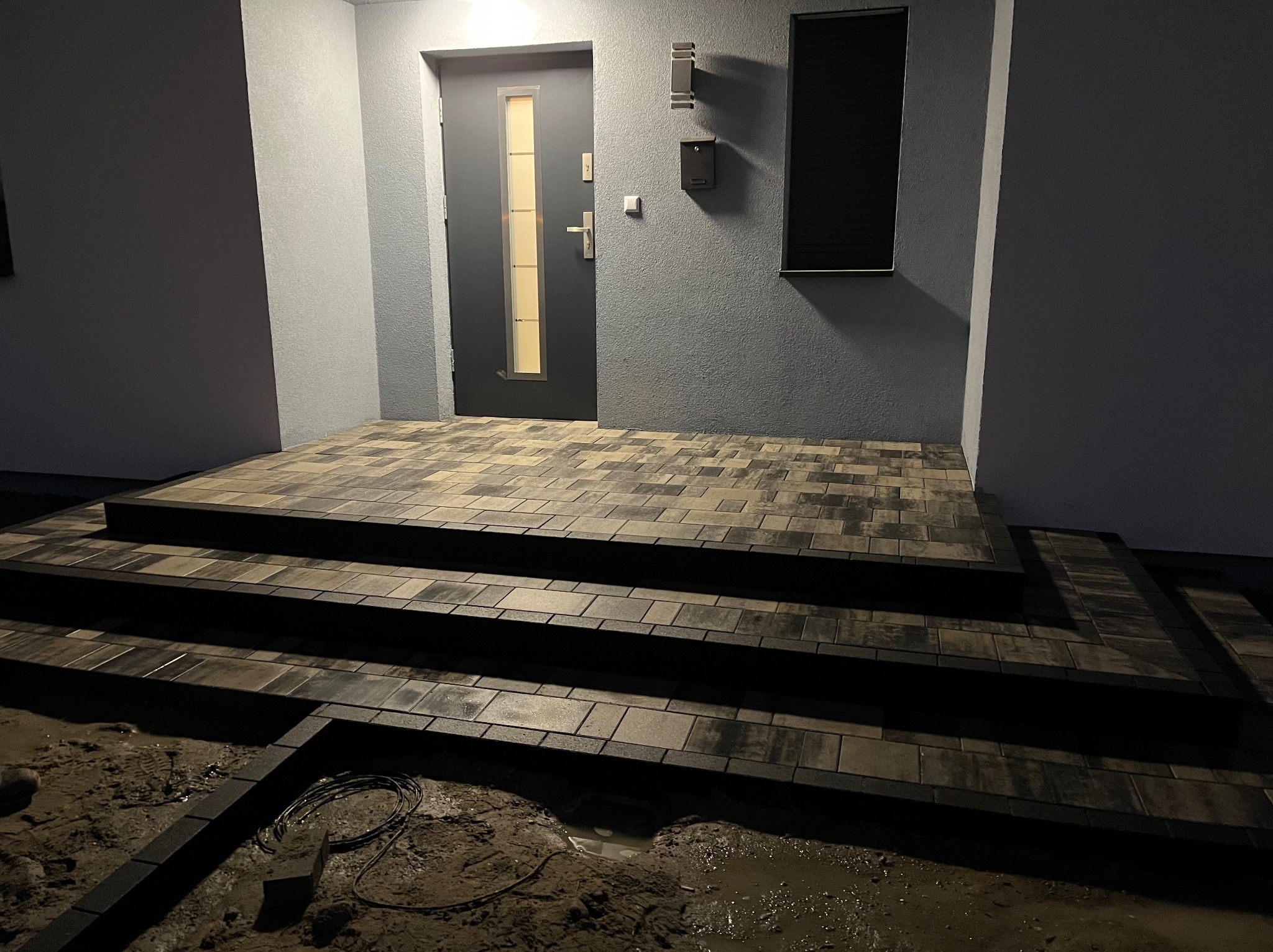 Schody wejściowe do domu wyłożone kostką brukową w odcieniach szarości i czerni, widoczne przewody elektryczne na ziemi, oświetlenie zewnętrzne, nowoczesna skrzynka na listy.