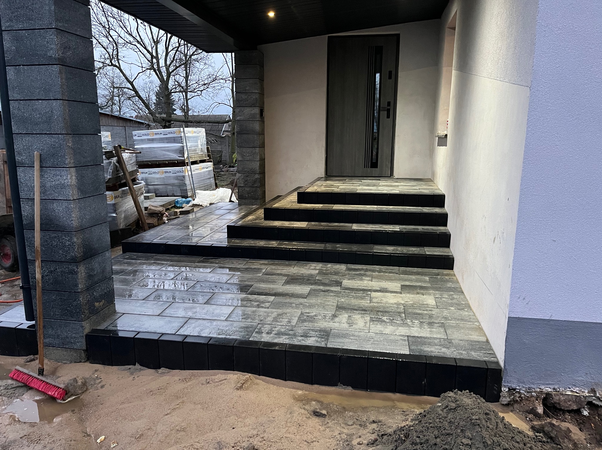 Nowoczesne wejście do domu wyłożone szarą kostką brukową z ciemnymi obrzeżami i schodami, widoczny stos kostki na paletach w tle oraz czerwona zmiotka przy kolumnie.