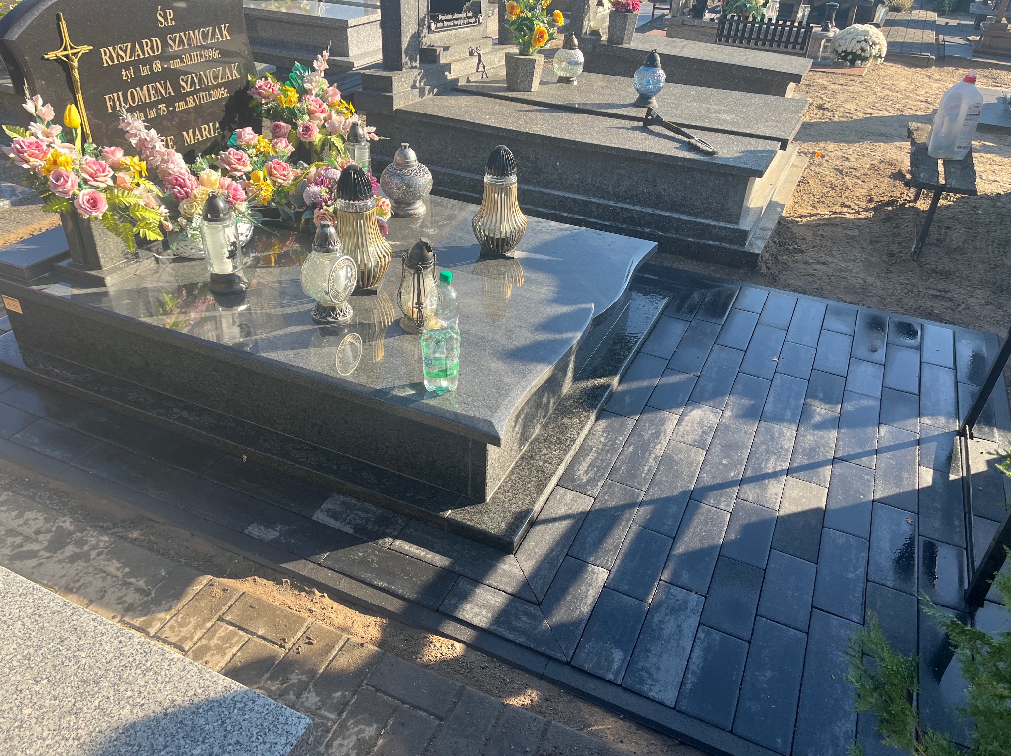 Ułożona kostka brukowa wokół nagrobka z ciemnego granitu, widoczne kwiaty i znicze na płycie nagrobnej, butelka wody, częściowo widoczna stara kostka brukowa na obrzeżach.