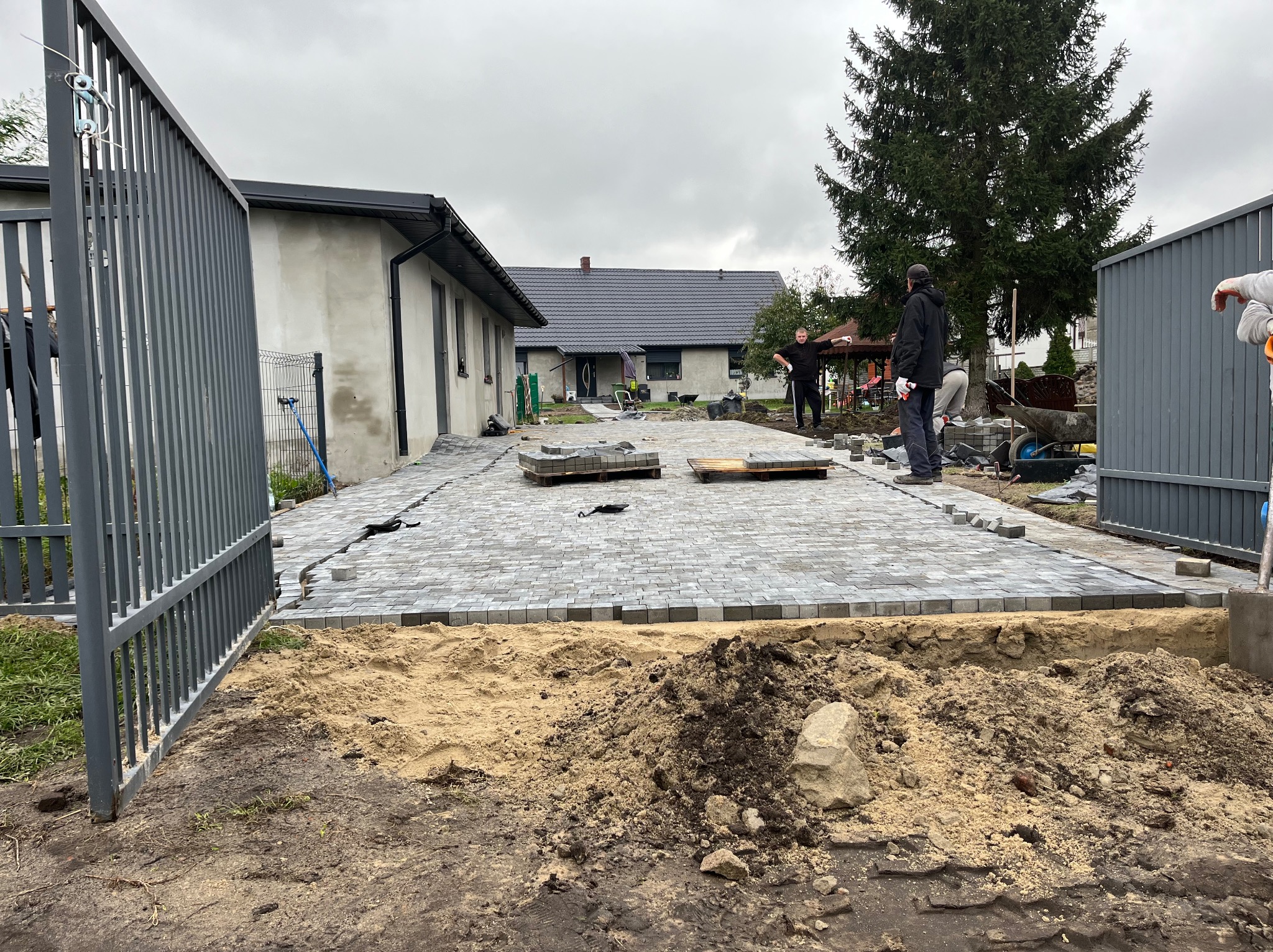 Kładzenie szarej kostki brukowej na podwórku domu jednorodzinnego, widoczni pracownicy w trakcie robót, palety z kostką i narzędzia, w tle drzewo iglaste i sąsiedni budynek.