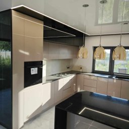 Domostan - Beżowa kuchnia z wyspą, oświetleniem LED i złotymi lampami wiszącymi, widziana pod kątem. Marmurowa podłoga i okno z widokiem na zieleń.