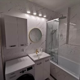 Domostan - Biała łazienka z marmurowymi płytkami, okrągłym lustrem w złotej ramie, białą szafką pod umywalką i pralką. Oświetlenie sufitowe.