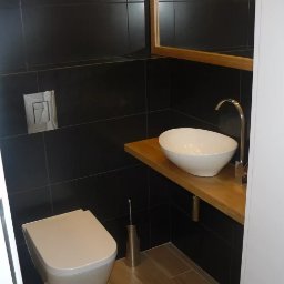 Domostan - Nowoczesna łazienka z czarnymi płytkami, drewnianym blatem pod umywalką w kształcie łzy i minimalistyczną toaletą. Lustro z drewnianą ramą nad umywalką.