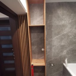 Domostan - Półka wnękowa z drewnianymi lamelami w łazience z szarymi płytkami. Czerwona poziomica oparta o bok. Widoczna wanna i bateria.