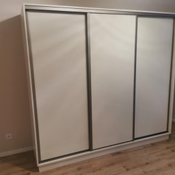 Domostan - Trzydrzwiowa szafa przesuwna w odcieniach bieli i szarości, dopasowana do wnętrza z panelami podłogowymi imitującymi drewno. Widoczny kontakt elektryczny na ścianie.