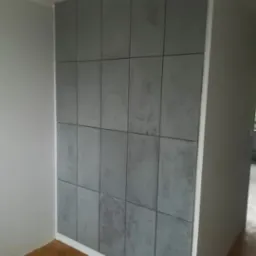 Ściana wykończona szarymi, kwadratowymi panelami imitującymi beton, z delikatną fakturą i widocznymi fugami, w jasnym wnętrzu. Minimalistyczny design.