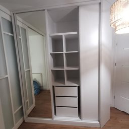 Domostan - Biała szafa przesuwna z lustrem i półkami, widok z boku. Na podłodze panele imitujące drewno. W tle widoczny fragment drzwi i lampy.