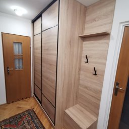 Domostan - Komplet mebli do przedpokoju z szafą przesuwną, siedziskiem i wieszakami, w odcieniach drewna, na tle parkietu i dywanu w orientalny wzór.