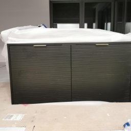 Domostan - Ciemna szafka z lamelowymi drzwiami, częściowo owinięta folią ochronną, stoi na kartonie w jasnym pomieszczeniu z dużymi oknami.