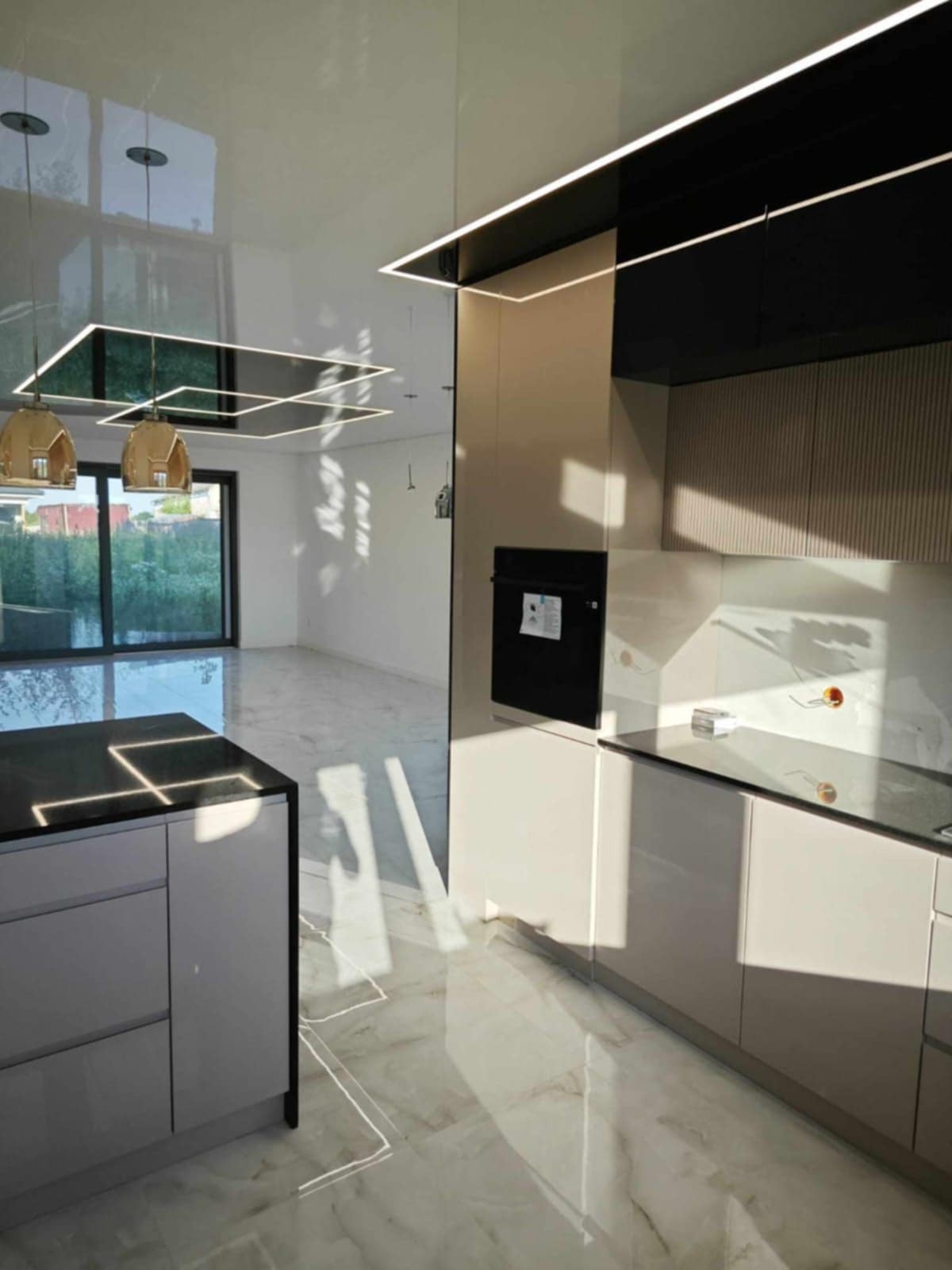 Nowoczesna kuchnia z połyskiem: beżowe szafki, czarny blat z oświetleniem LED, marmurowa podłoga i odbicie okna w suficie.
