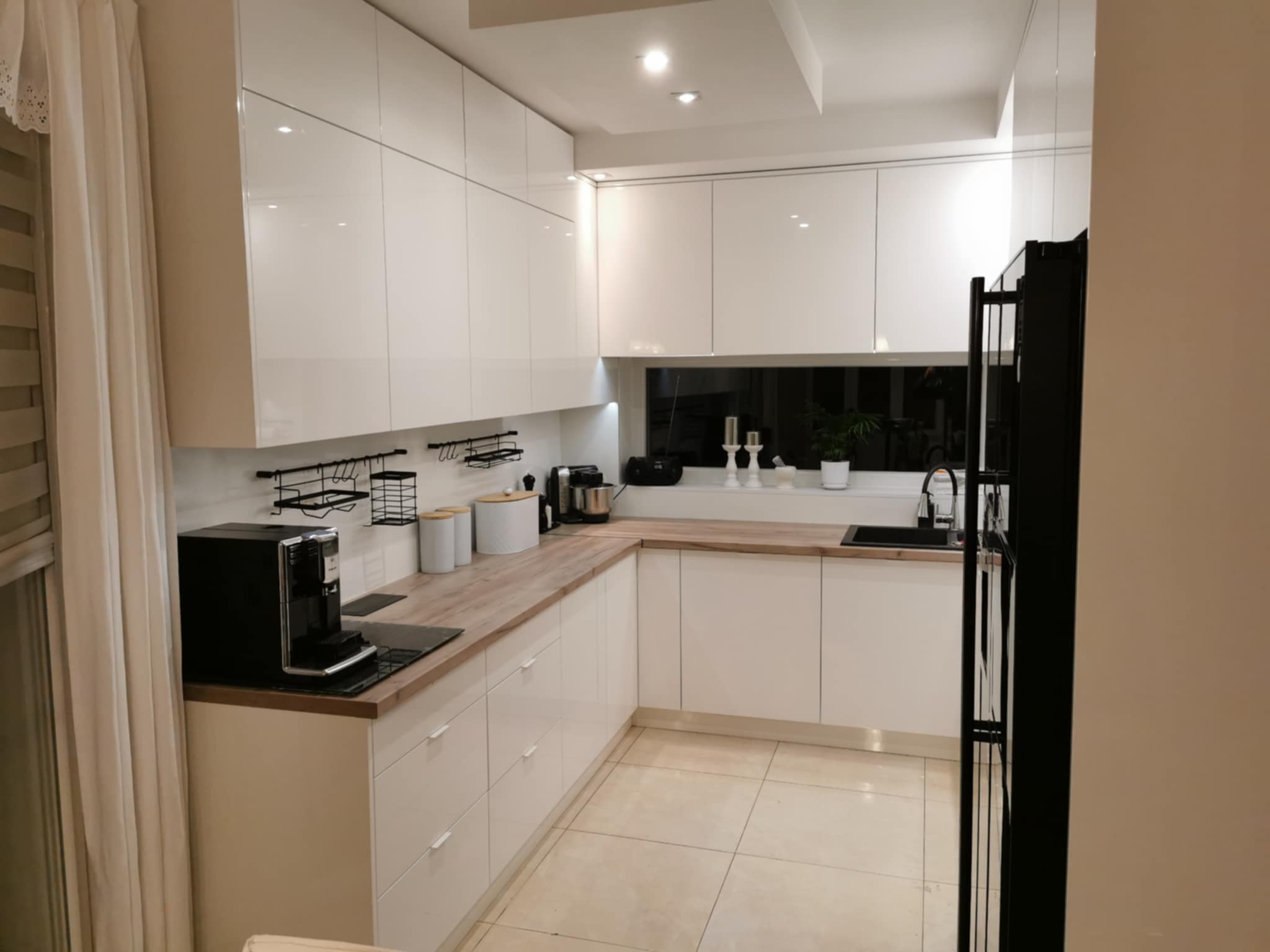 Jasna kuchnia z białymi, lakierowanymi szafkami, drewnianym blatem i czarnym sprzętem AGD. Okno z widokiem na zieleń. Minimalistyczny design, nowoczesne oświetlenie.