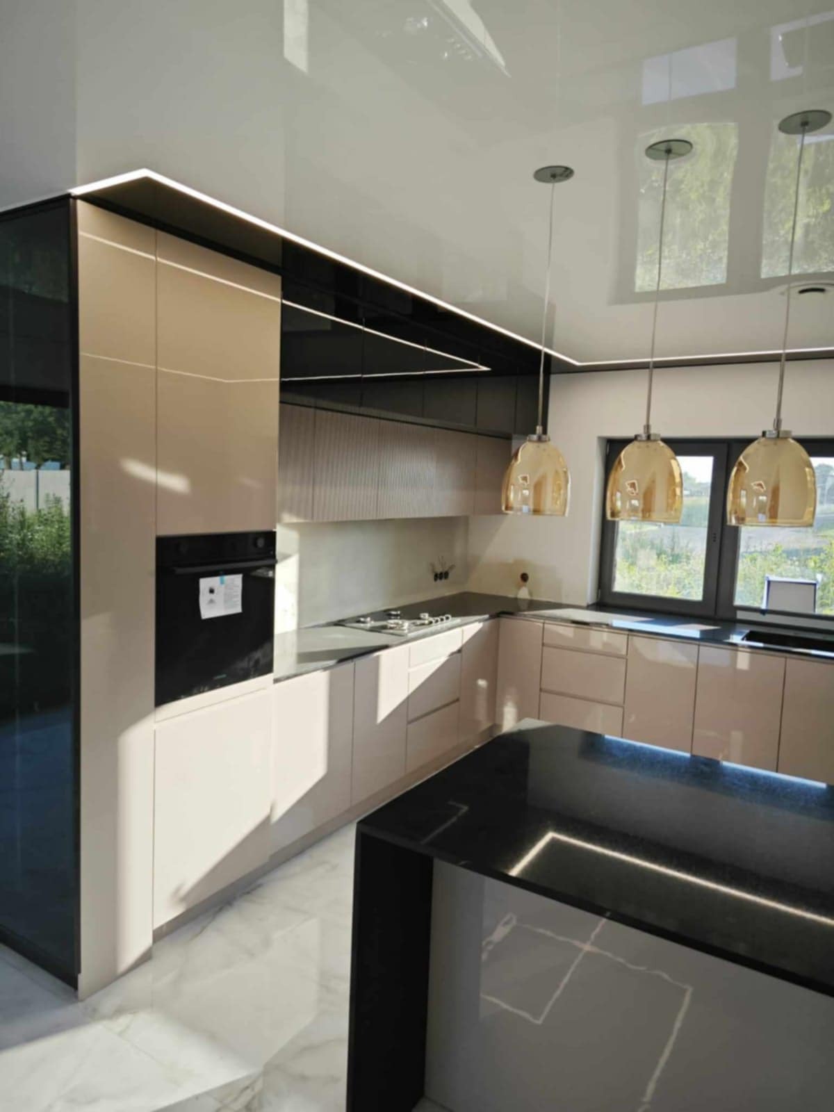 Beżowa kuchnia z wyspą, oświetleniem LED i złotymi lampami wiszącymi, widziana pod kątem. Marmurowa podłoga i okno z widokiem na zieleń.
