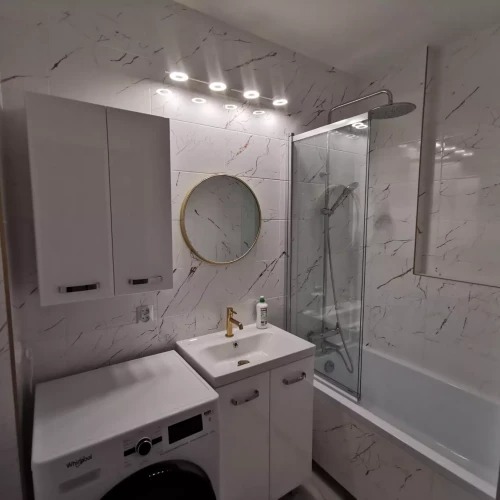 Biała łazienka z marmurowymi płytkami, okrągłym lustrem w złotej ramie, białą szafką pod umywalką i pralką. Oświetlenie sufitowe.