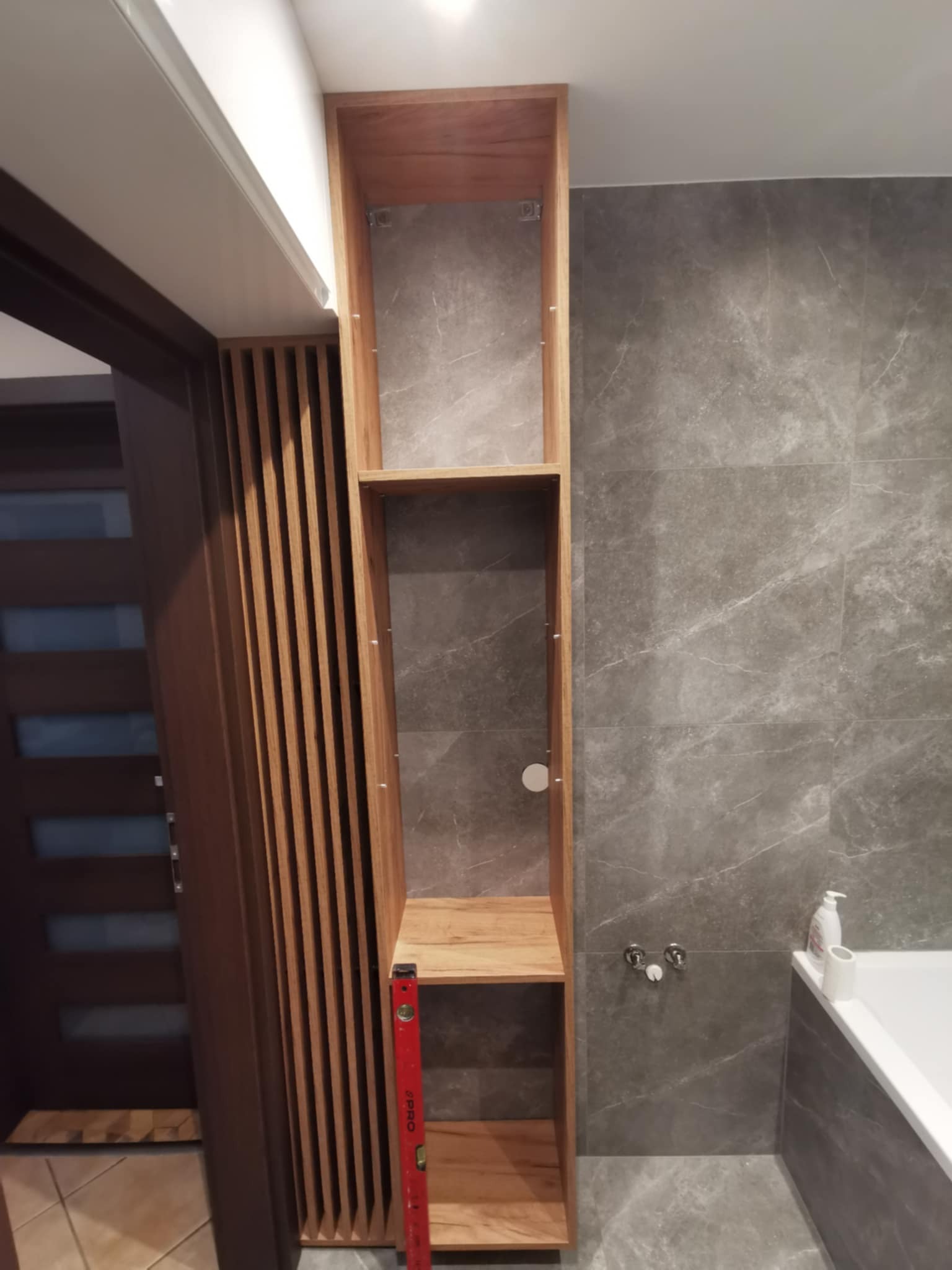 Półka wnękowa z drewnianymi lamelami w łazience z szarymi płytkami. Czerwona poziomica oparta o bok. Widoczna wanna i bateria.
