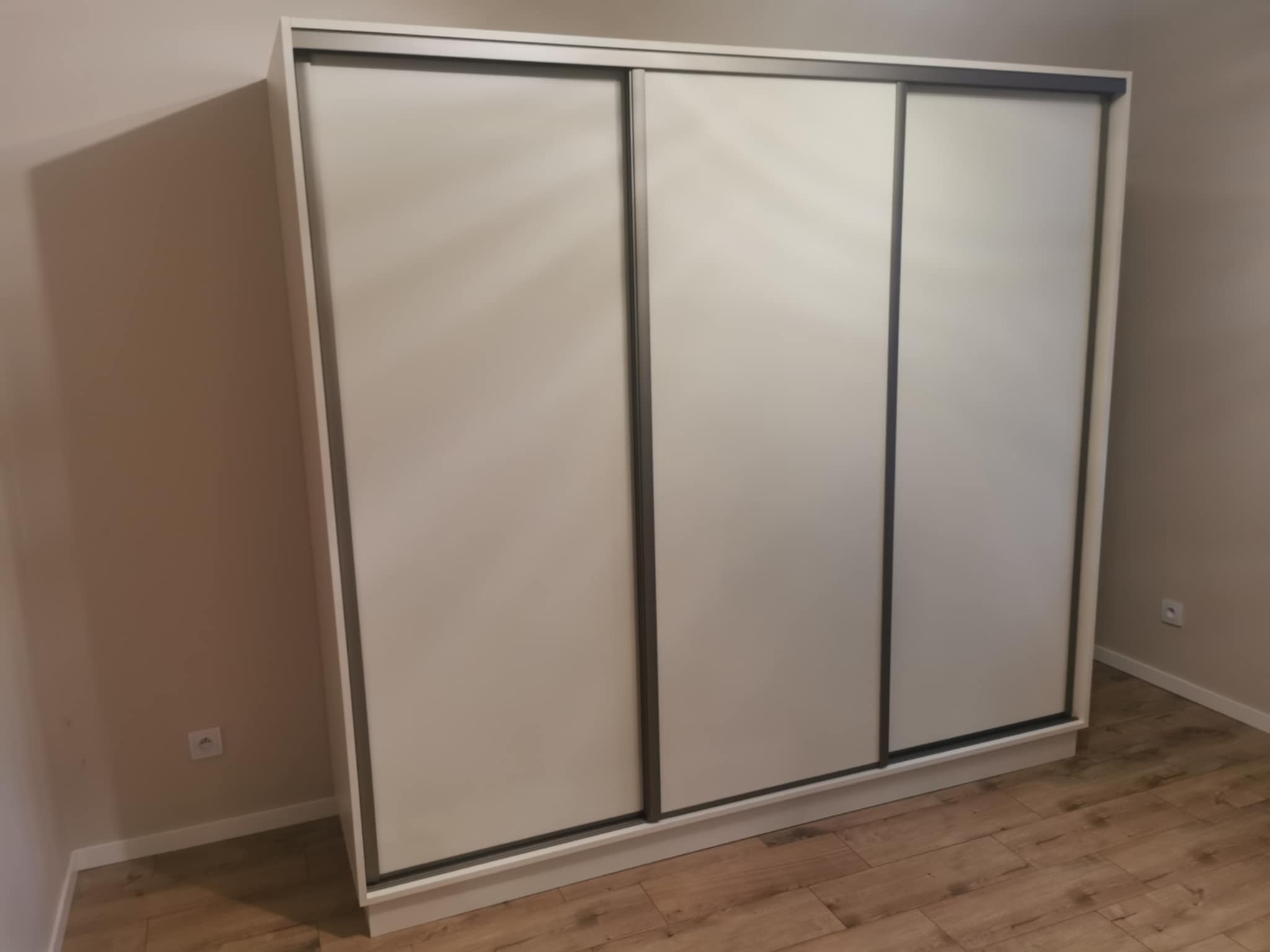 Trzydrzwiowa szafa przesuwna w odcieniach bieli i szarości, dopasowana do wnętrza z panelami podłogowymi imitującymi drewno. Widoczny kontakt elektryczny na ścianie.