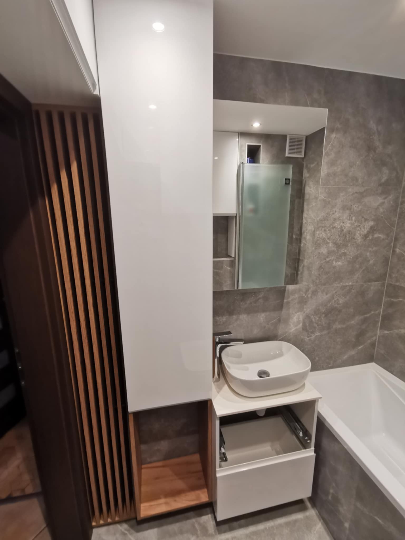 Nowoczesna łazienka z marmurowymi płytkami, białą umywalką nablatową, szafką z drewnianymi akcentami i lustrem. Minimalistyczny design z otwartą szufladą.
