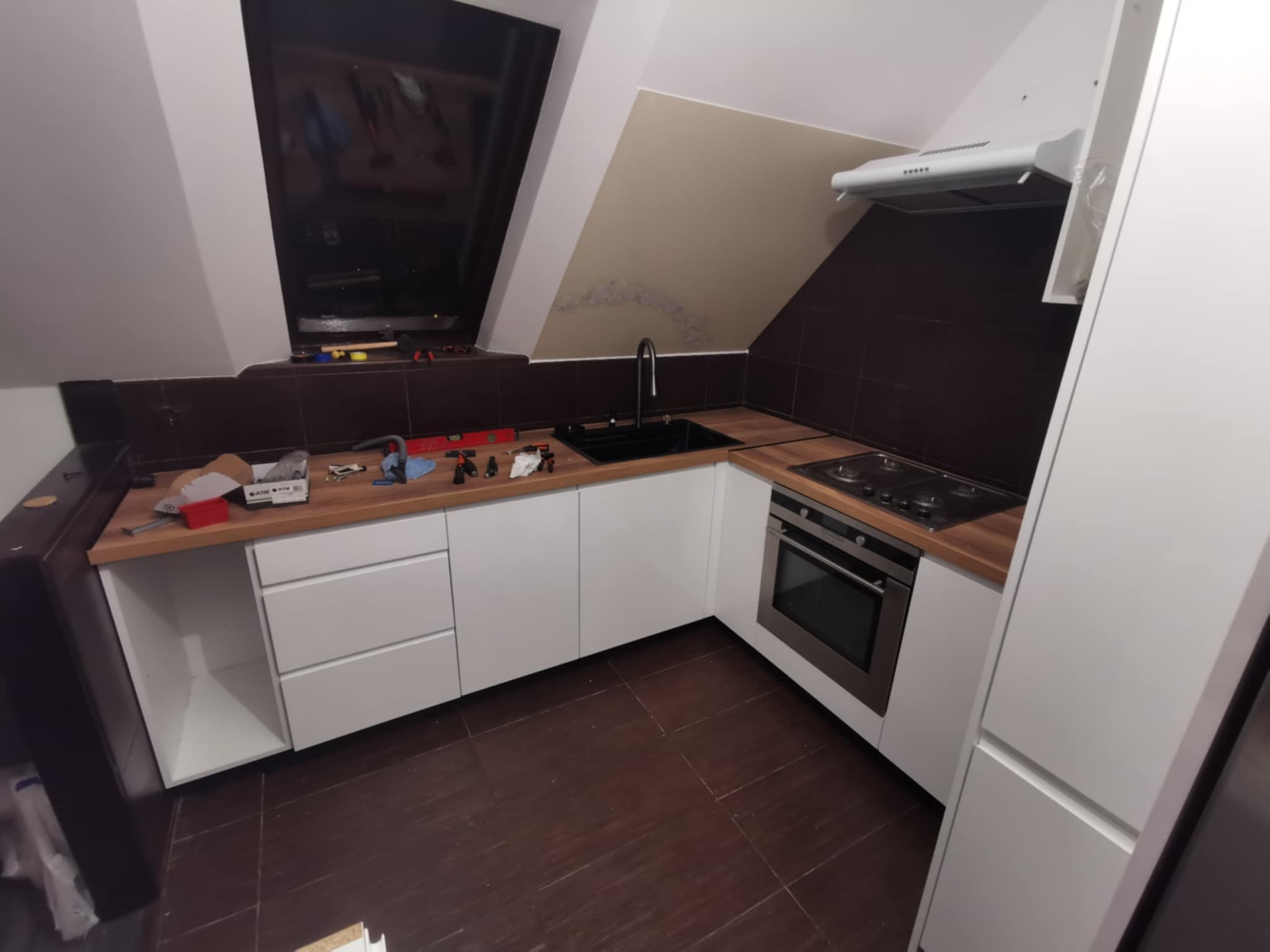 Nowoczesna kuchnia na poddaszu z białymi szafkami, drewnianym blatem i czarnym zlewem. Widoczne narzędzia sugerują etap montażu.