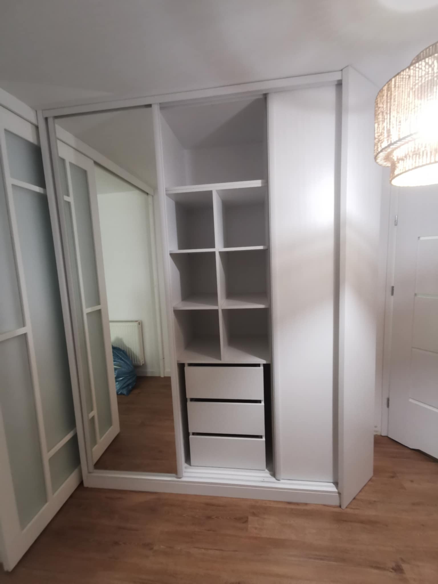 Biała szafa przesuwna z lustrem i półkami, widok z boku. Na podłodze panele imitujące drewno. W tle widoczny fragment drzwi i lampy.