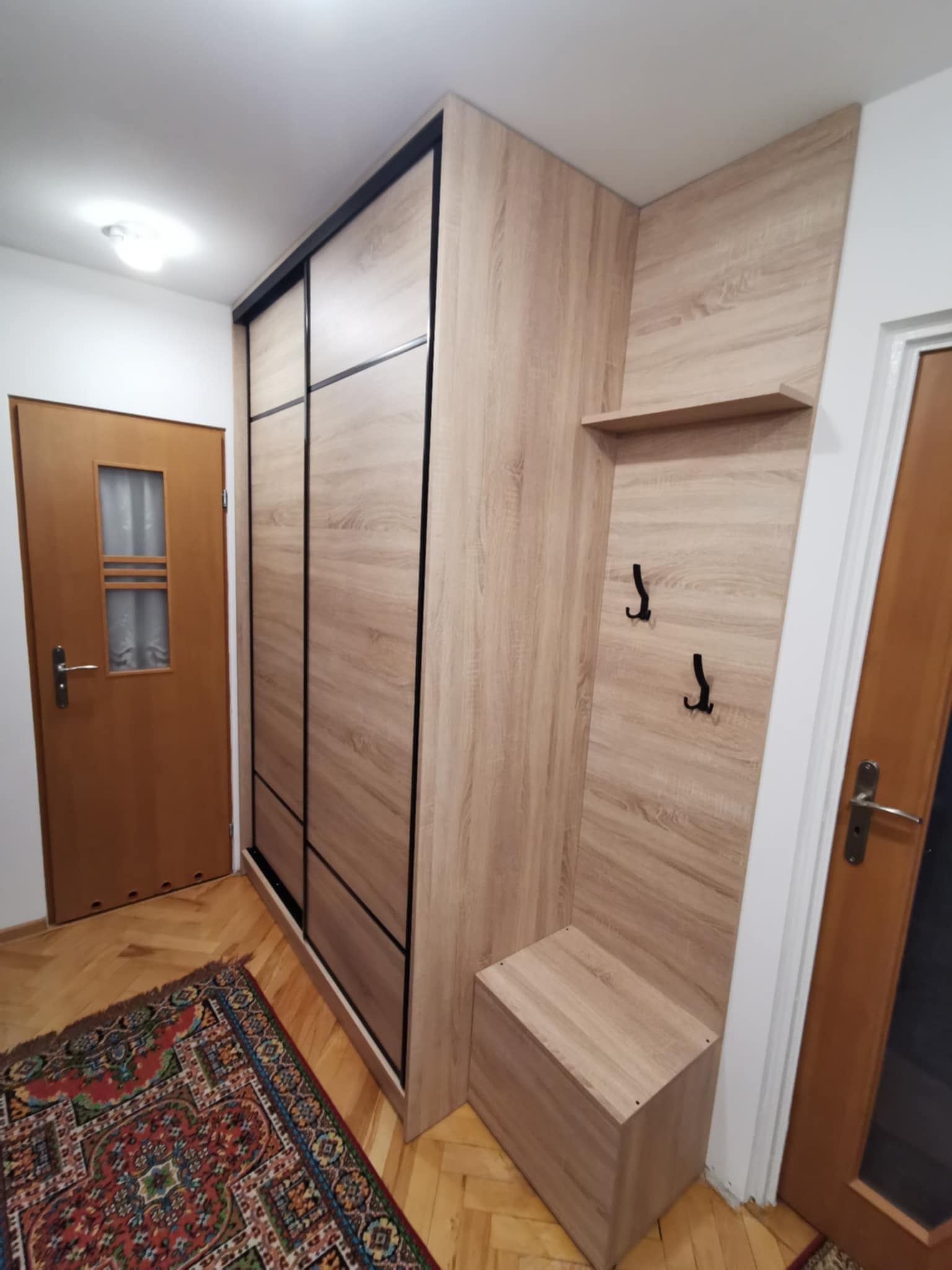 Komplet mebli do przedpokoju z szafą przesuwną, siedziskiem i wieszakami, w odcieniach drewna, na tle parkietu i dywanu w orientalny wzór.