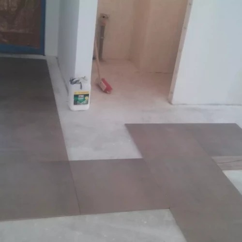 Układanie paneli podłogowych w jasnym wnętrzu. Widoczna chemia budowlana przy ścianie i fragment czerwonej szczotki. Częściowo ułożone panele w szarym odcieniu.